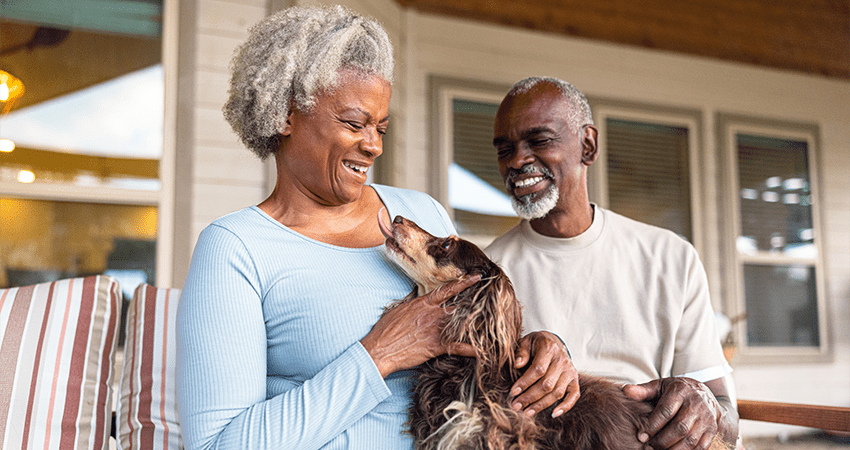 An older Black man and woman sit on a porch holding their dog